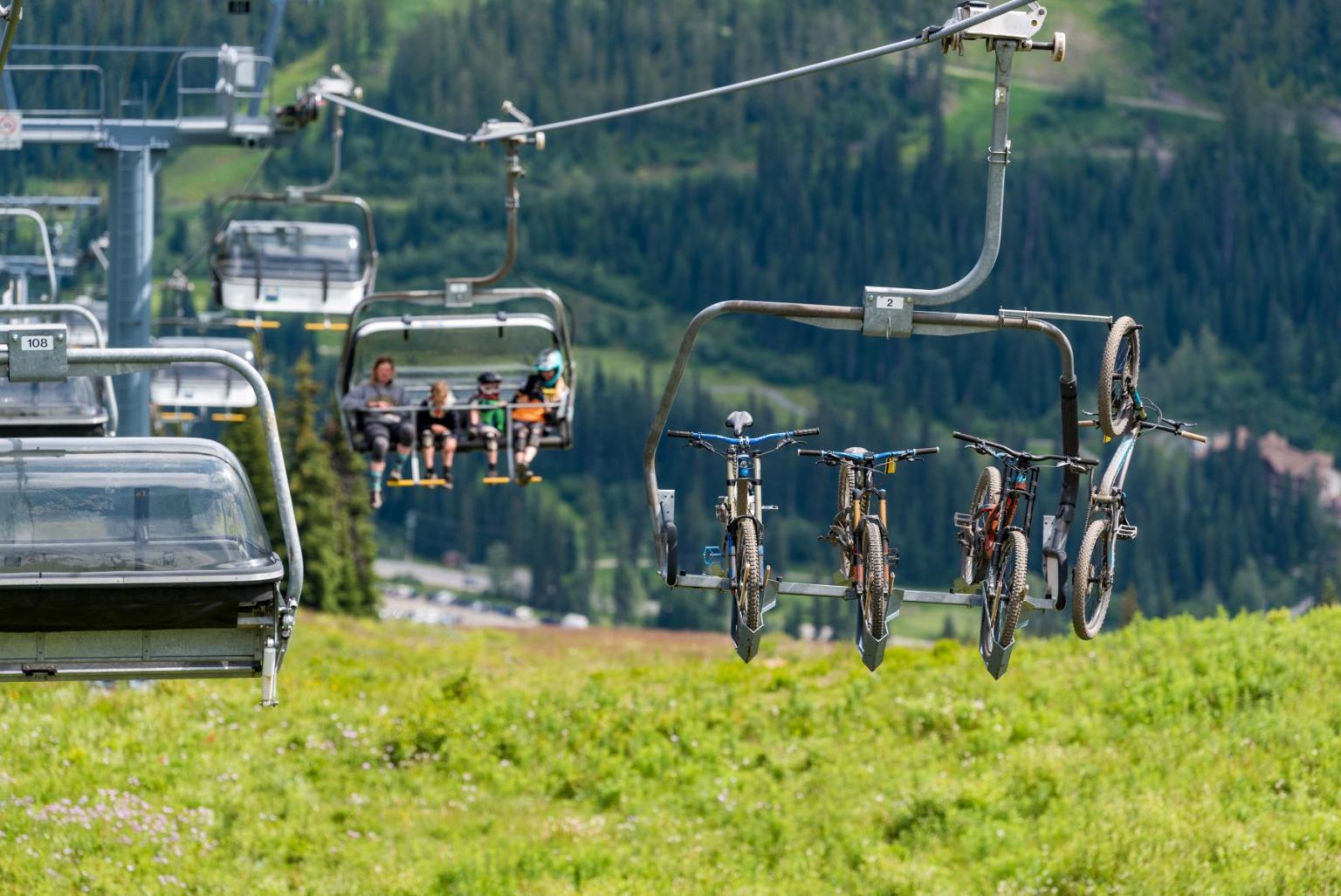 Ski lift carrying people and bicycles over green Mountainside.