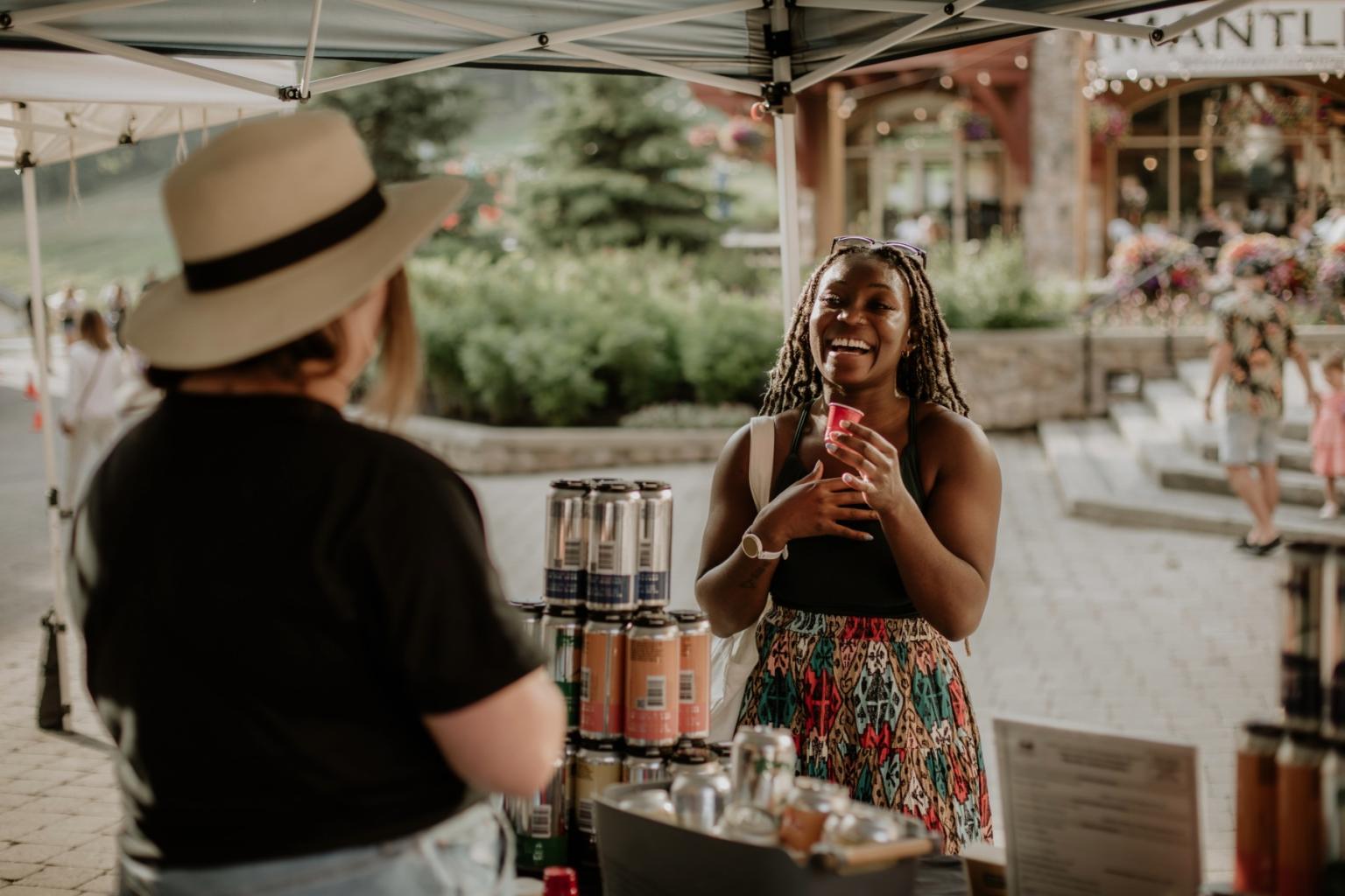 Two women at an outdoor market stand, one smiling with a drink.