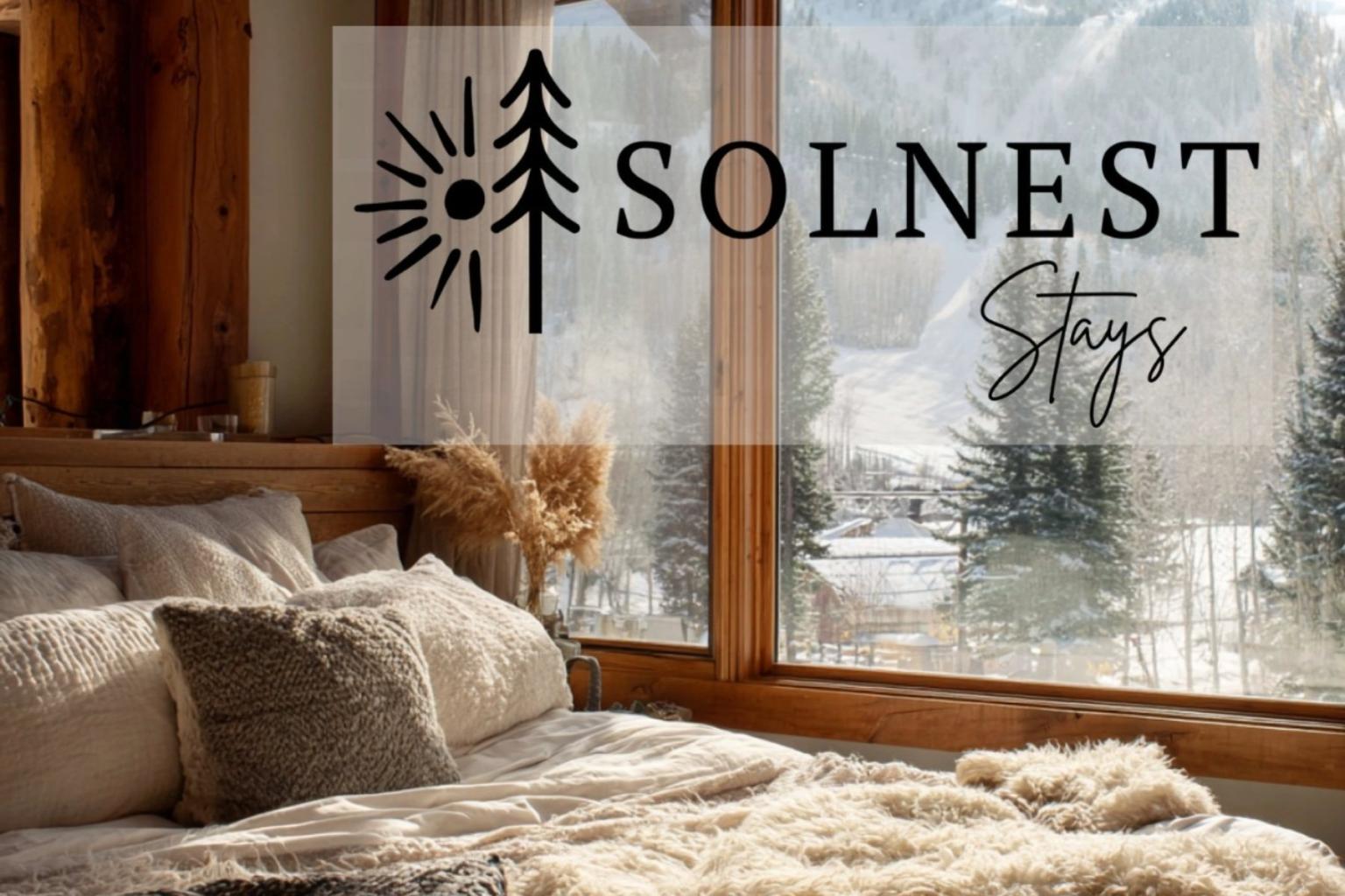 Solnest Stays - Cozy bedroom with plush pillows and a snowy mountain view through large windows.