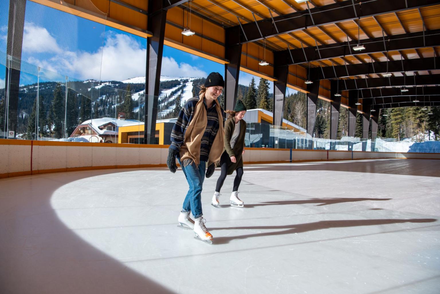 Two people ice skating in a sunny, indoor rink with mountain views.