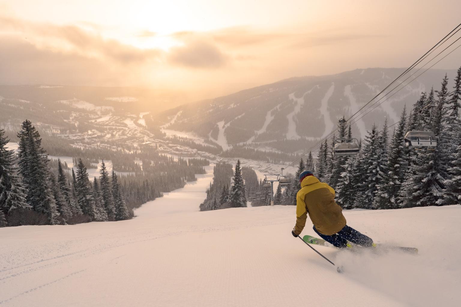 Skier on snowy slope at sunrise, mountains in background.