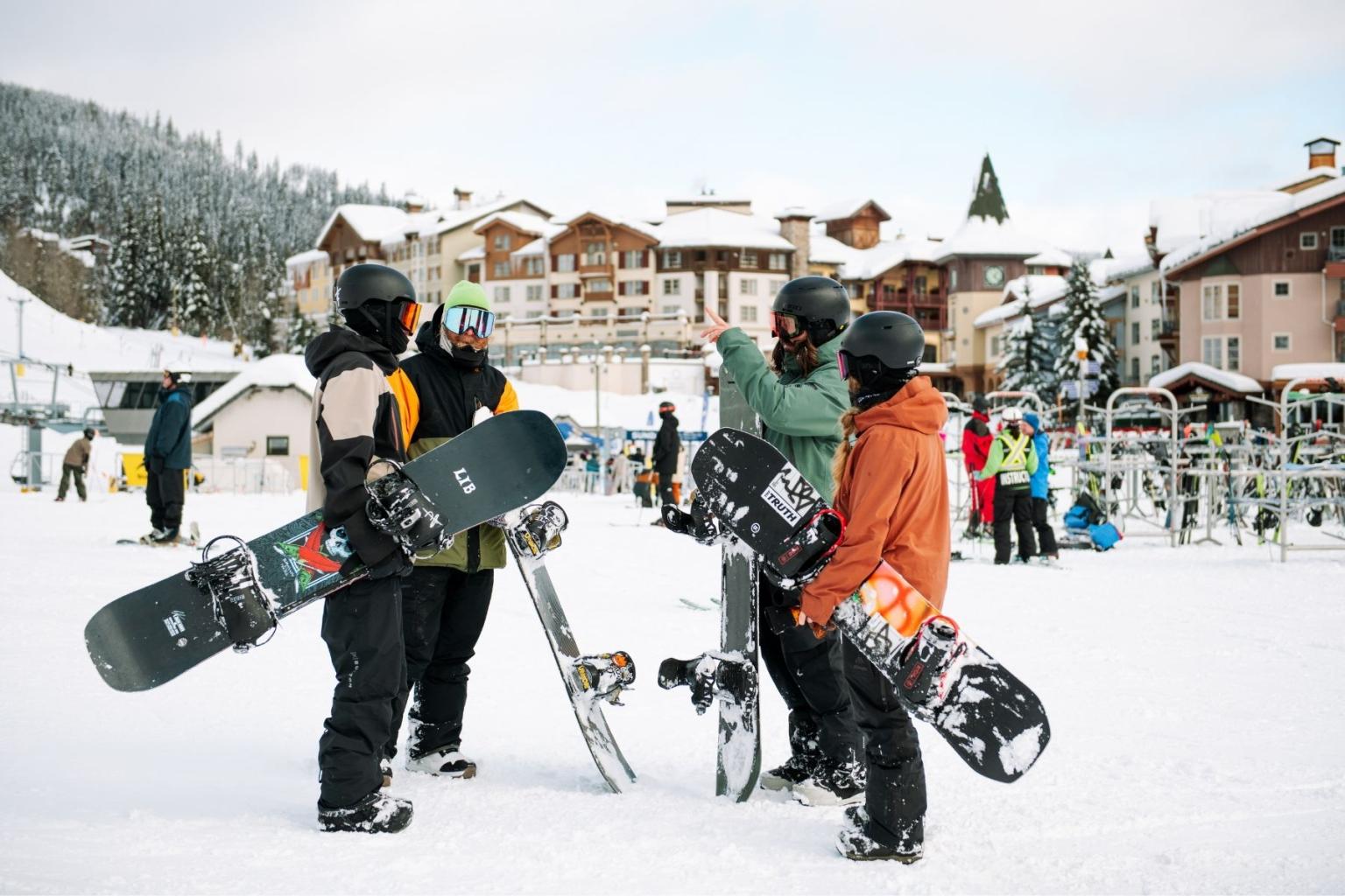 Four snowboarders chatting on a snowy slope near mountain lodges.
