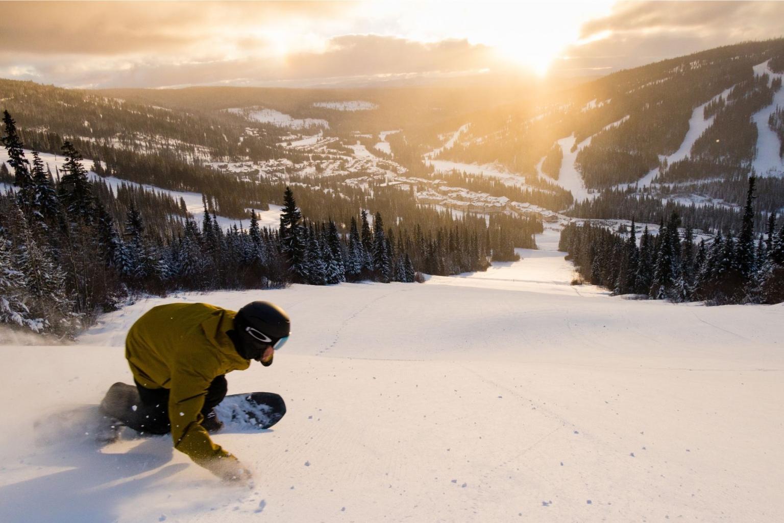 Snowboarder on a snowy slope at sunrise, surrounded by mountains and trees.