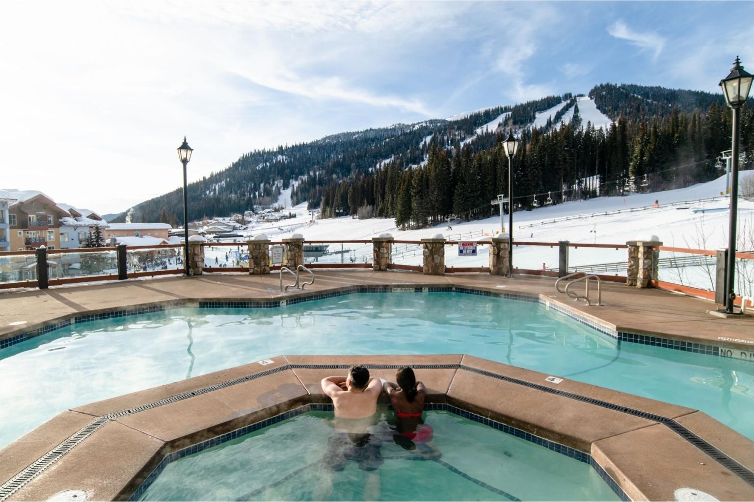 Two people relax in an outdoor hot tub, snowy mountains in the background.