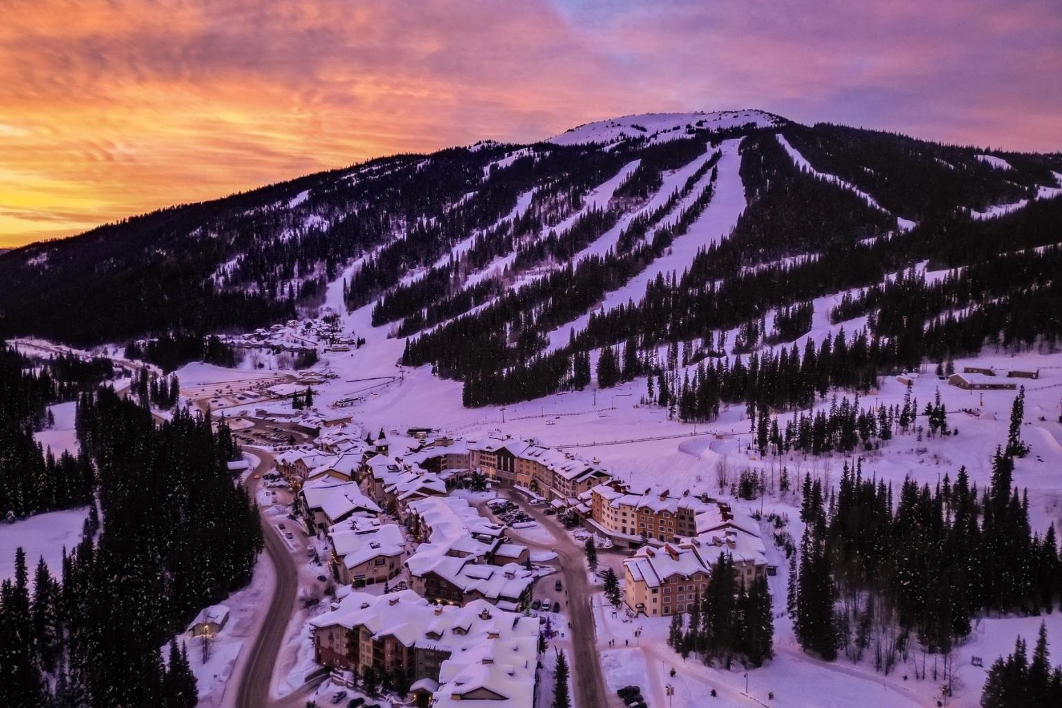 Snowy mountain village at sunset with ski slopes, pine trees, and colorful sky.