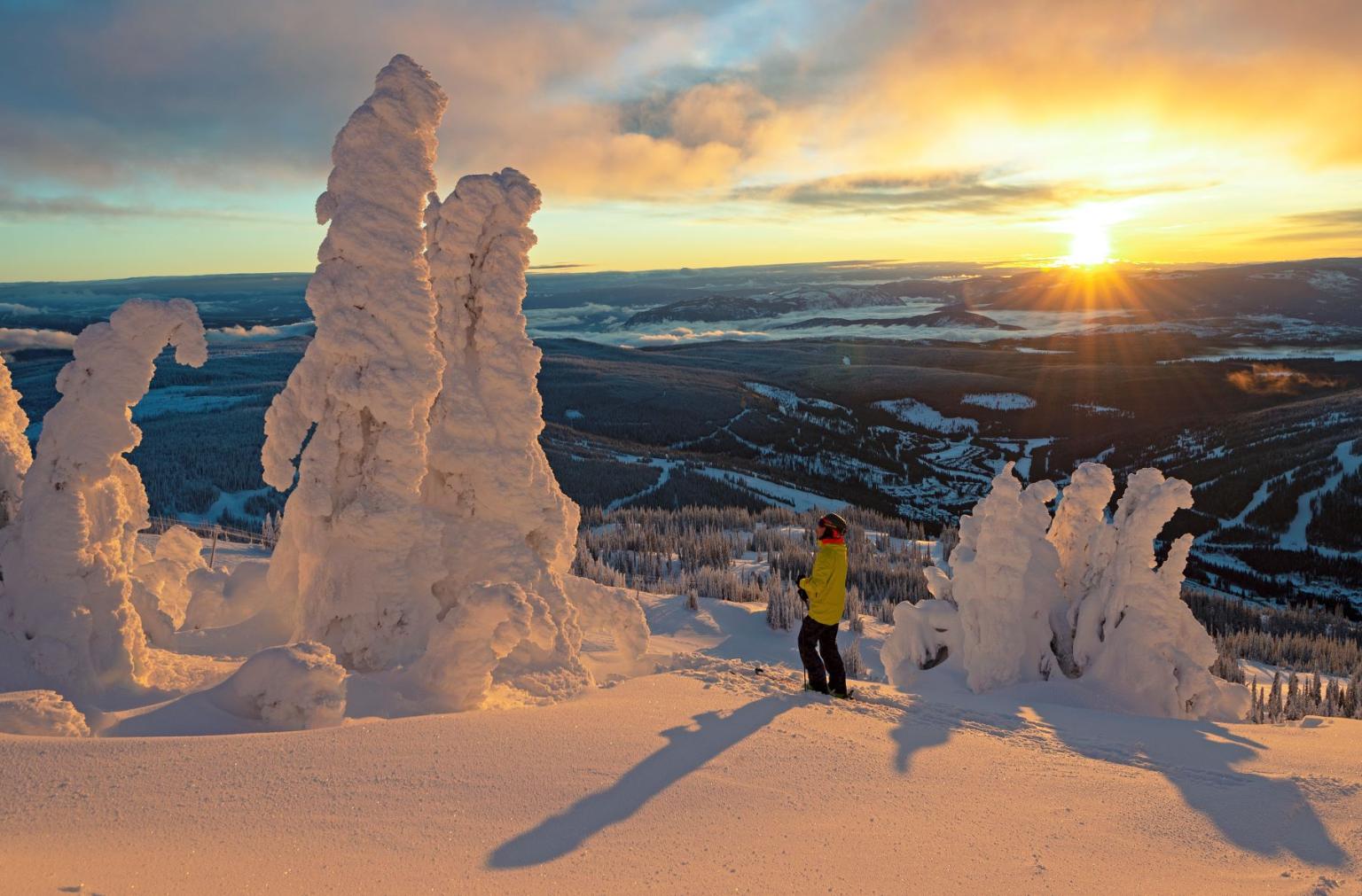 Snow-covered landscape at sunset, person in yellow jacket admiring view.