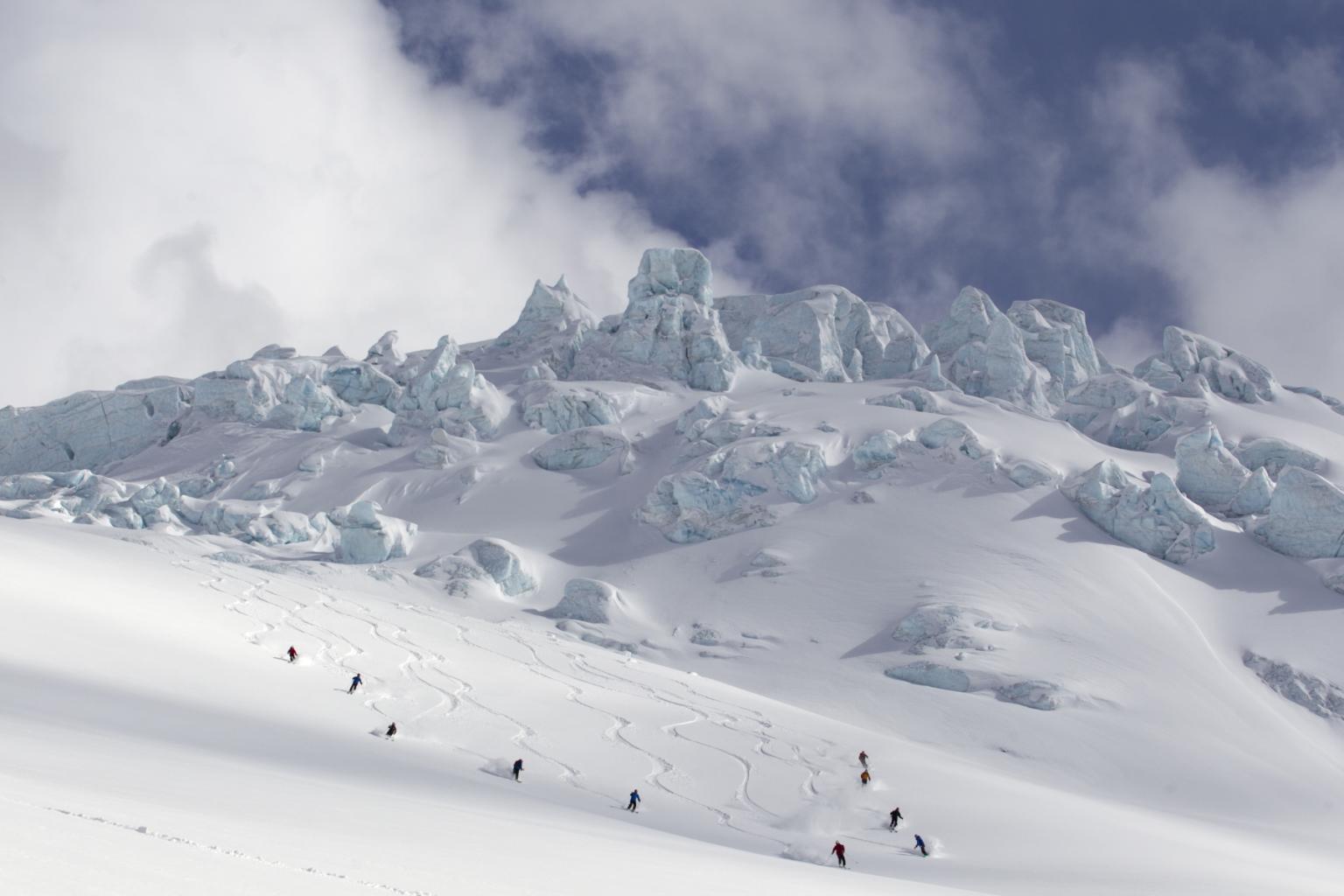 Skiers descending a snowy mountain slope beneath a cloudy sky.