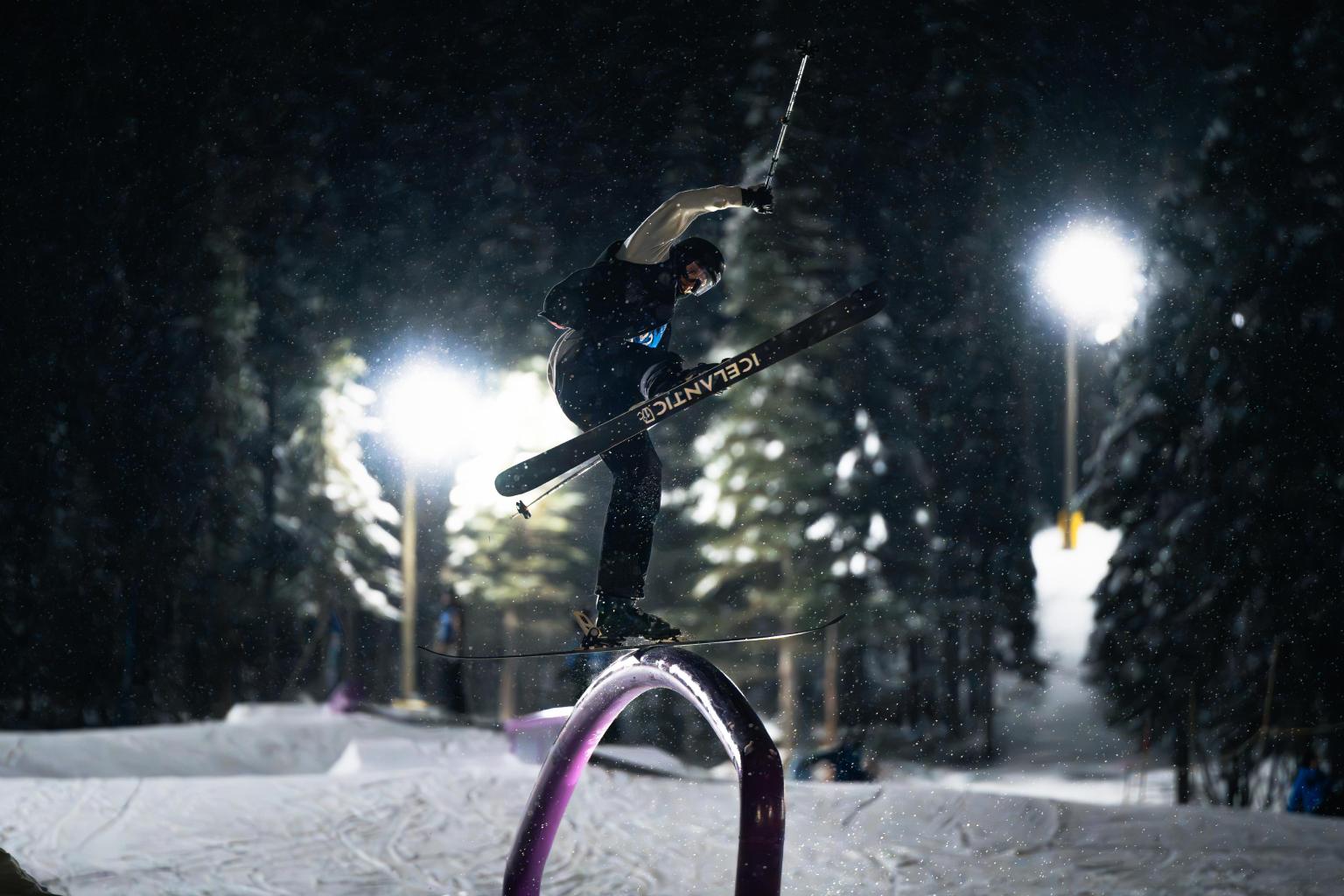Skier performing a jump on a rail at night, surrounded by snowy trees.