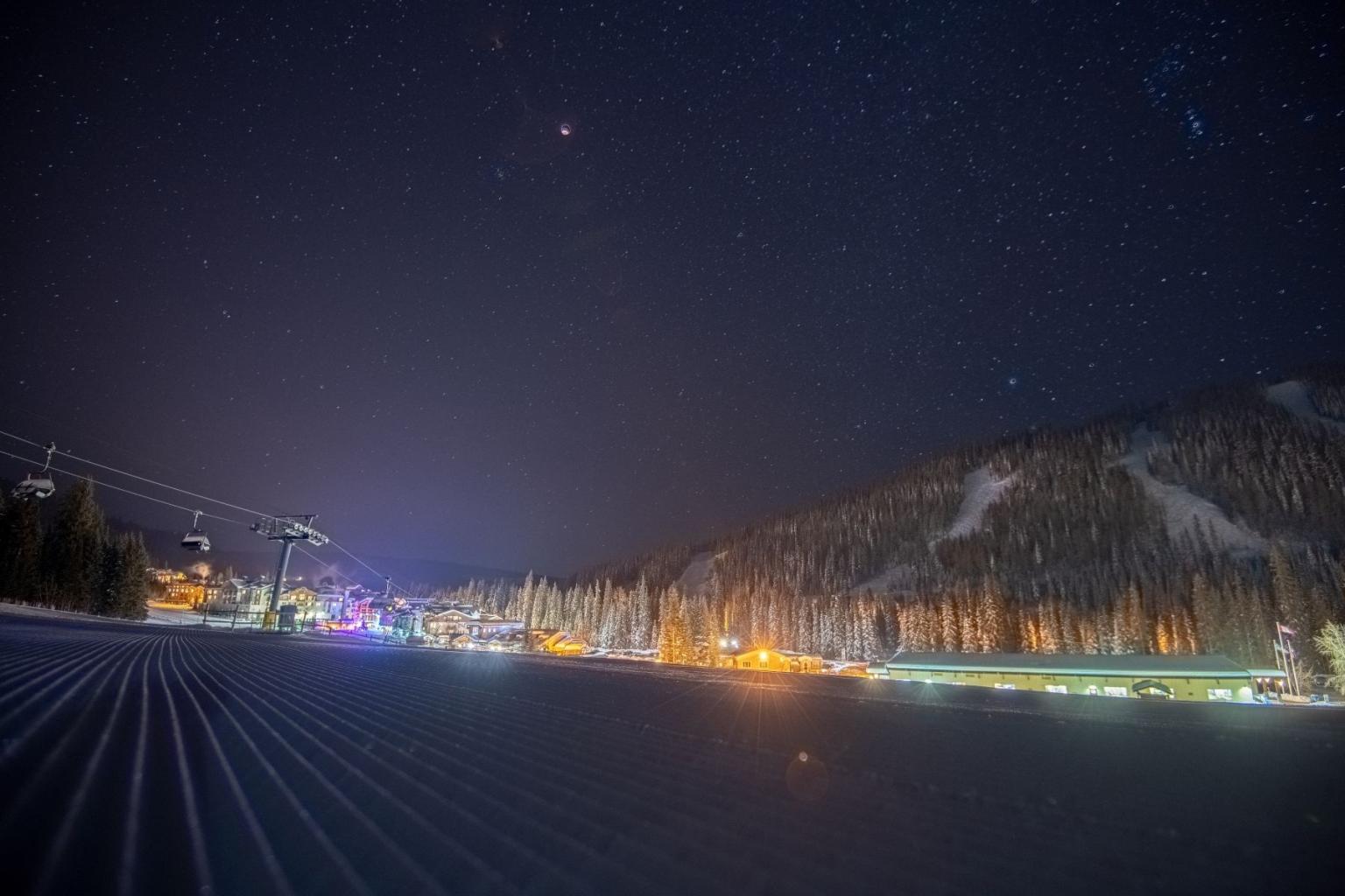 Starry night over snowy ski resort with lights and mountains.