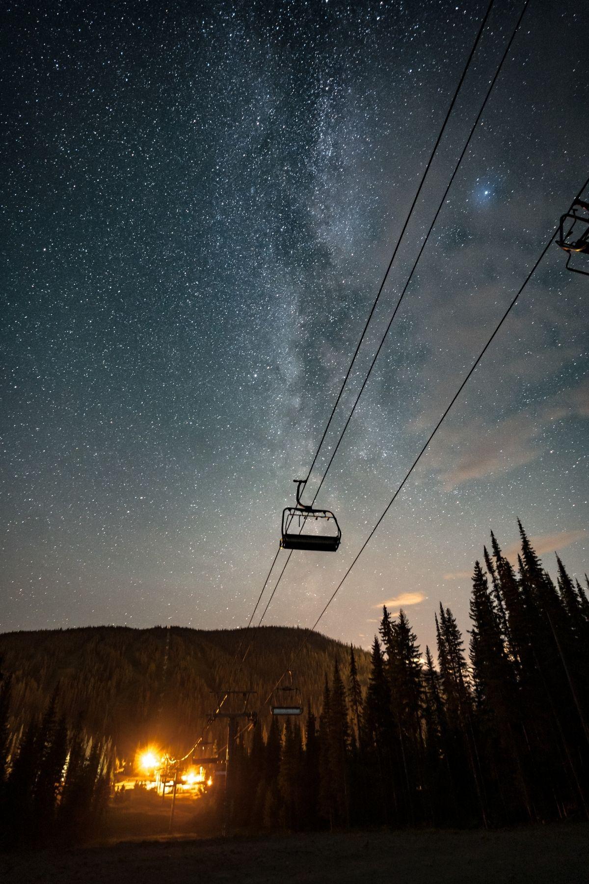 Starry sky over a mountain with a silhouetted ski lift and glowing lights below.