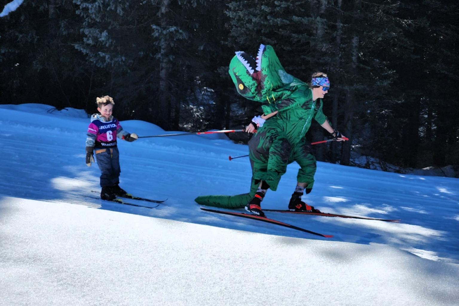 Child skiing behind person in dinosaur costume on snowy slope.