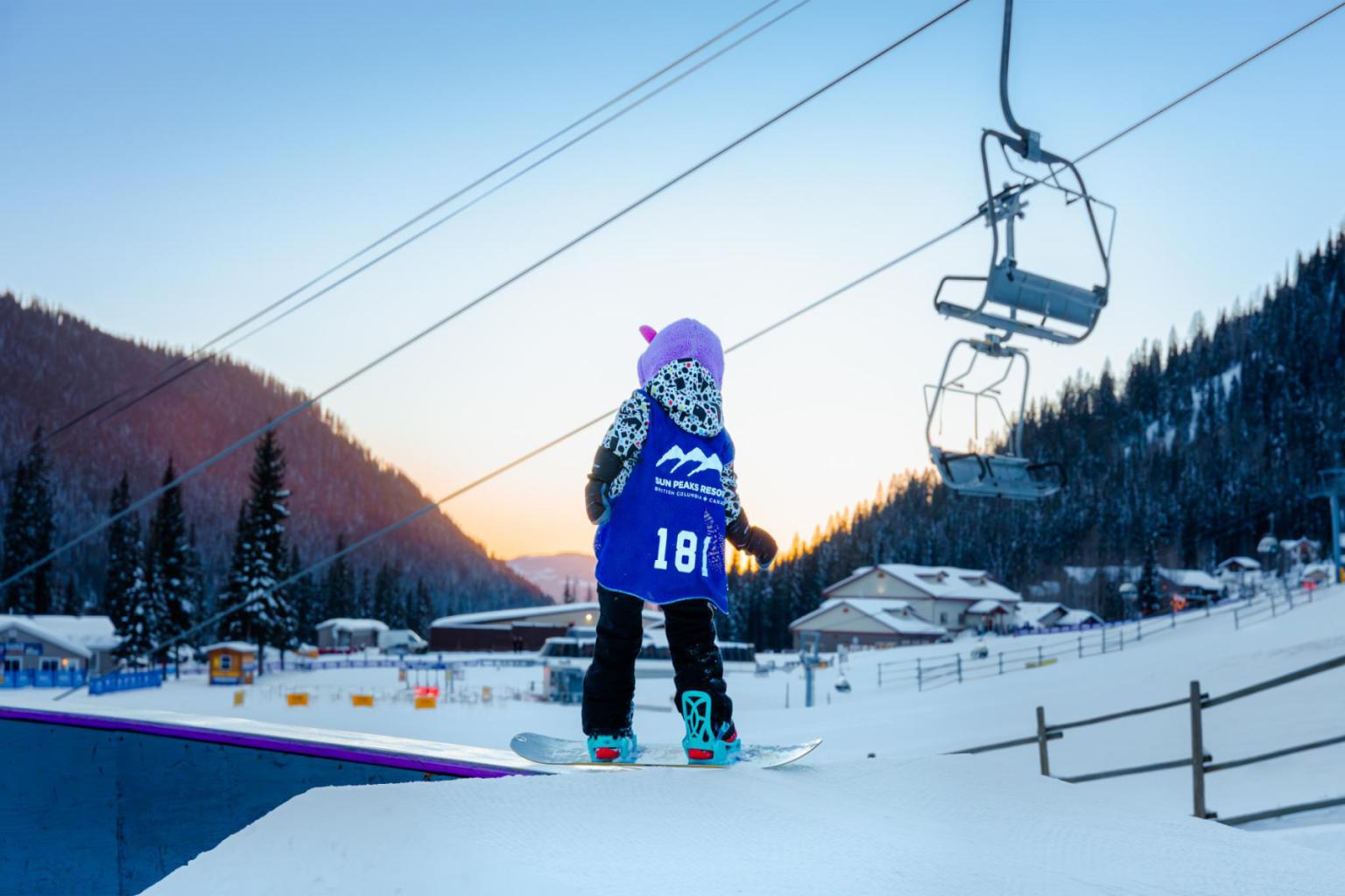 Child snowboarder in purple helmet on snowy slope near ski lift at dawn.