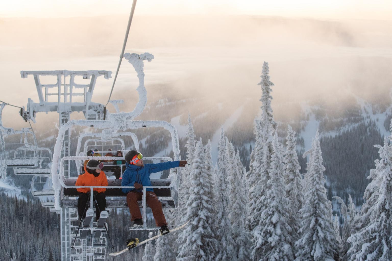 Skiers on a snowy chairlift ride through frosty trees at sunrise.
