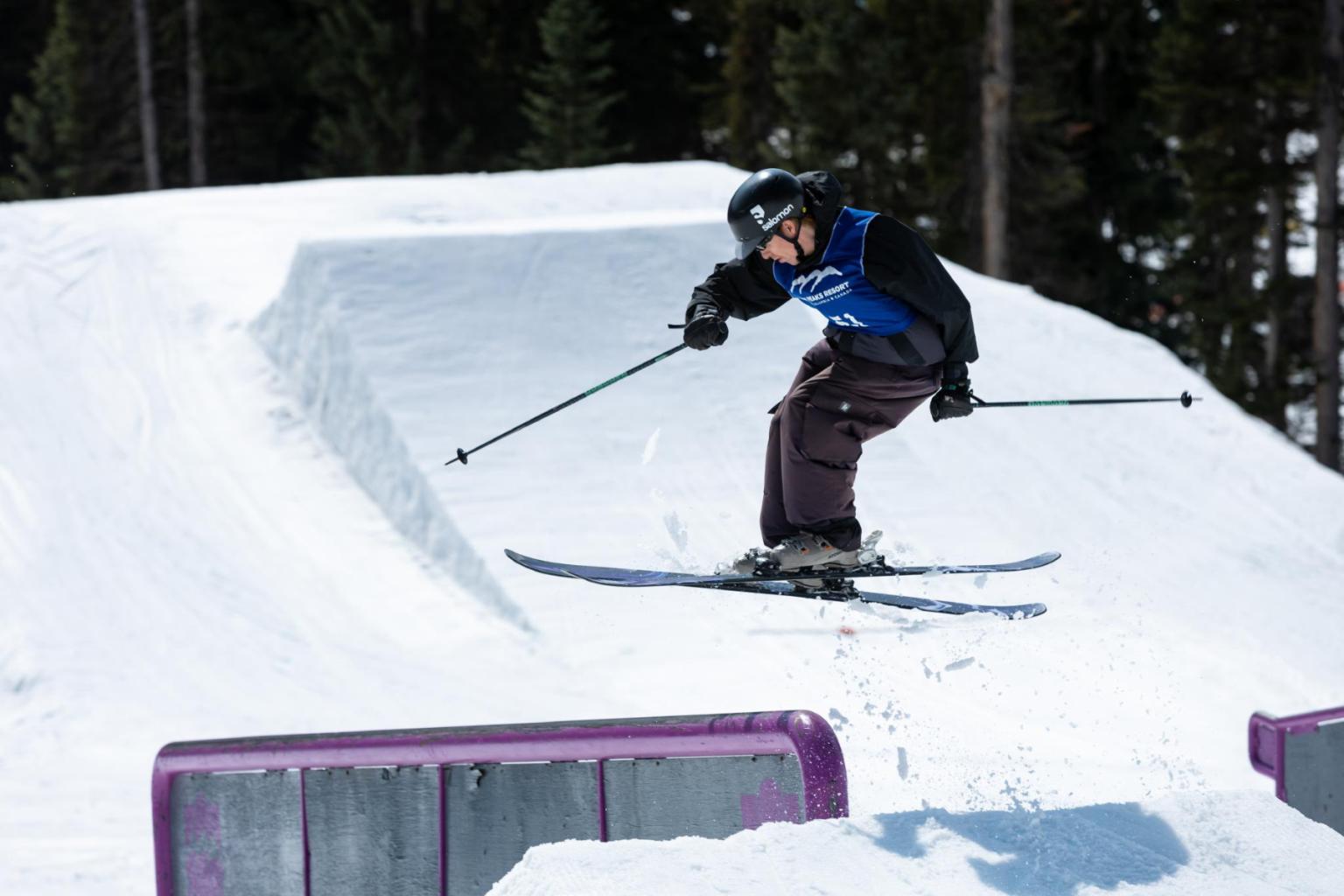 Skier in mid-air jump over snowy ramp, wearing helmet and blue vest.