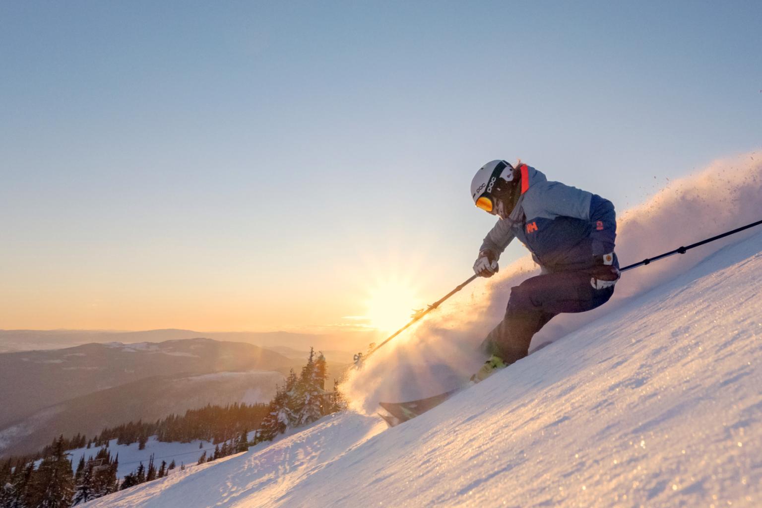 Skier on snowy slope at sunset, emitting spray under a clear sky.
