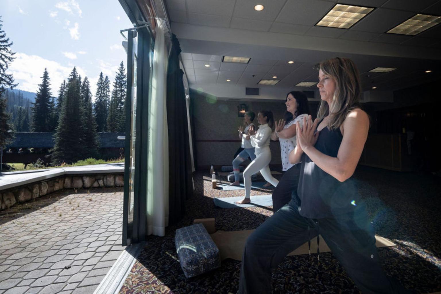 Women practicing yoga indoors with a view of trees outside.
