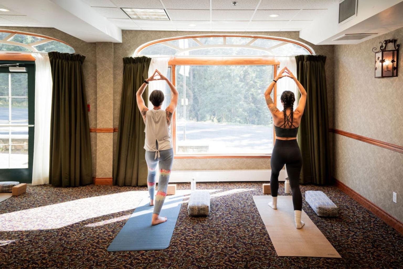 Two people practicing yoga indoors near a large sunny window.