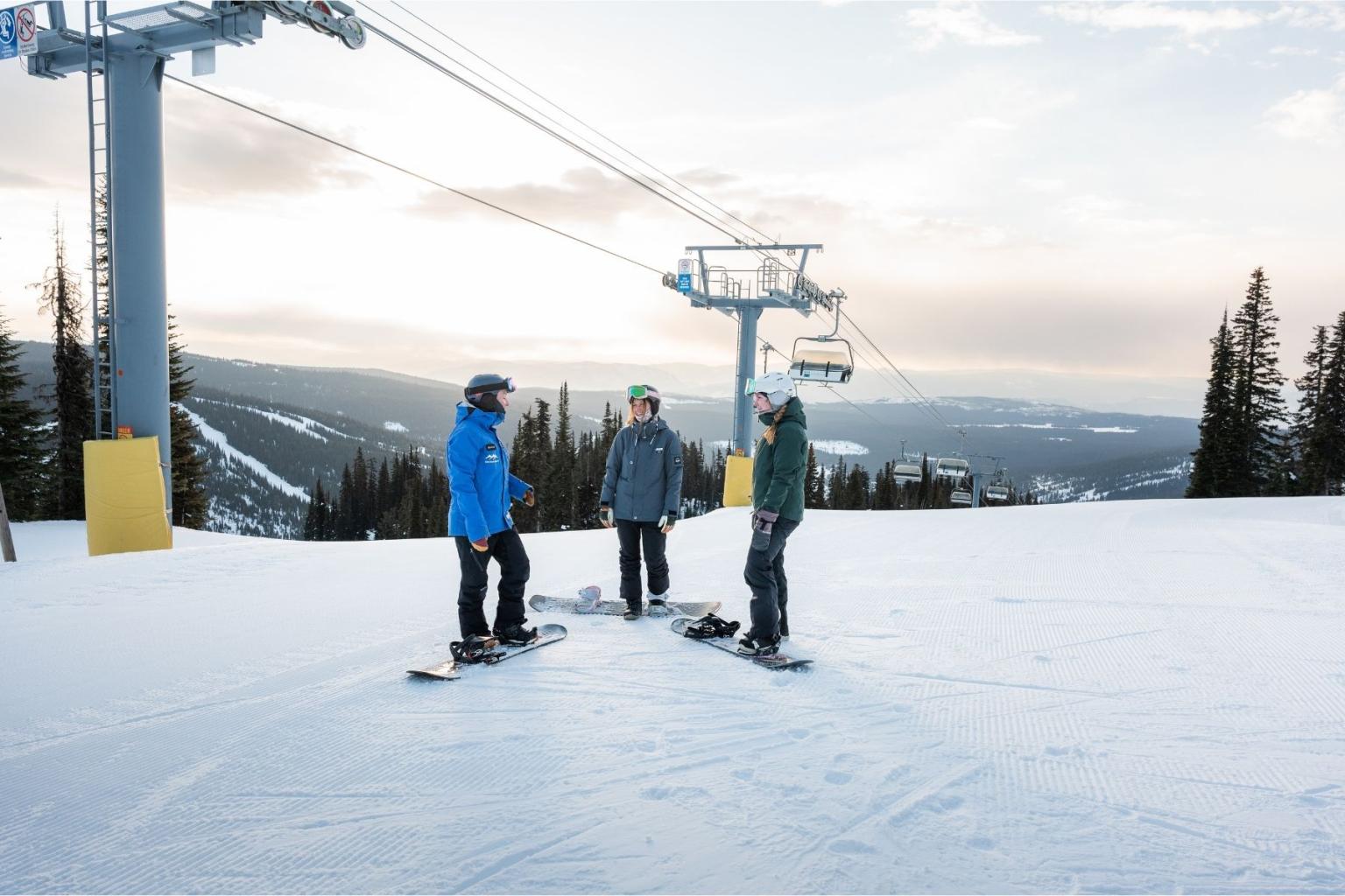 Three people with snowboards stand on a snowy slope near a ski lift.
