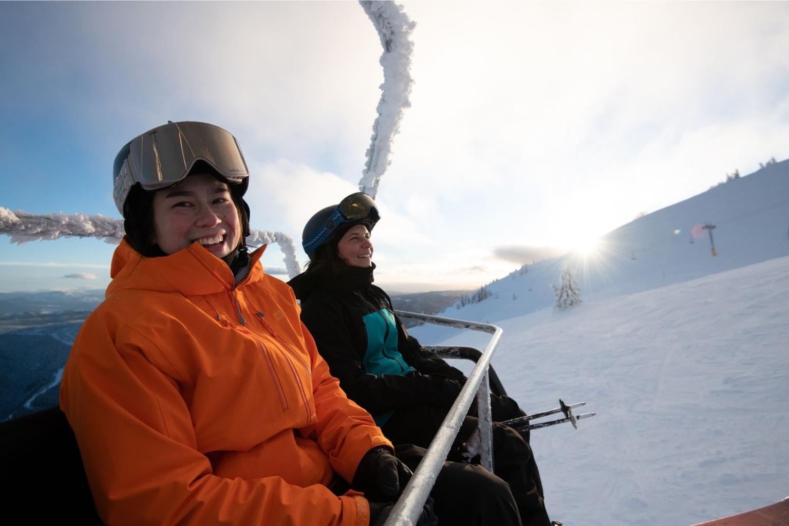 Skiers in bright jackets ride a snowy chairlift at sunrise.
