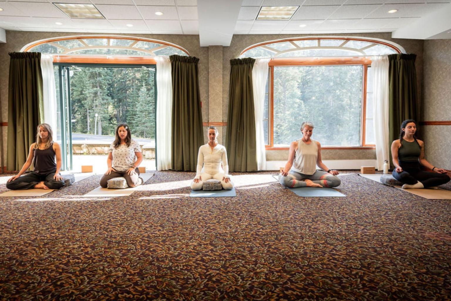 Yoga class with five people meditating indoors, sunlight through large windows.