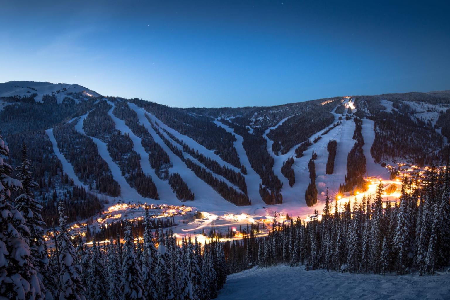 Snowy ski resort at night, slopes illuminated, surrounded by trees under a twilight sky.