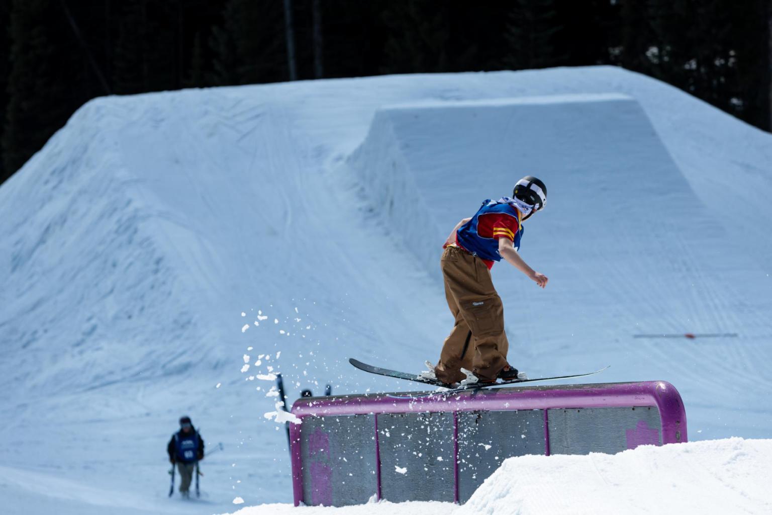 Snowboarder balancing on a rail in a snowy park, with a large ramp in the background.