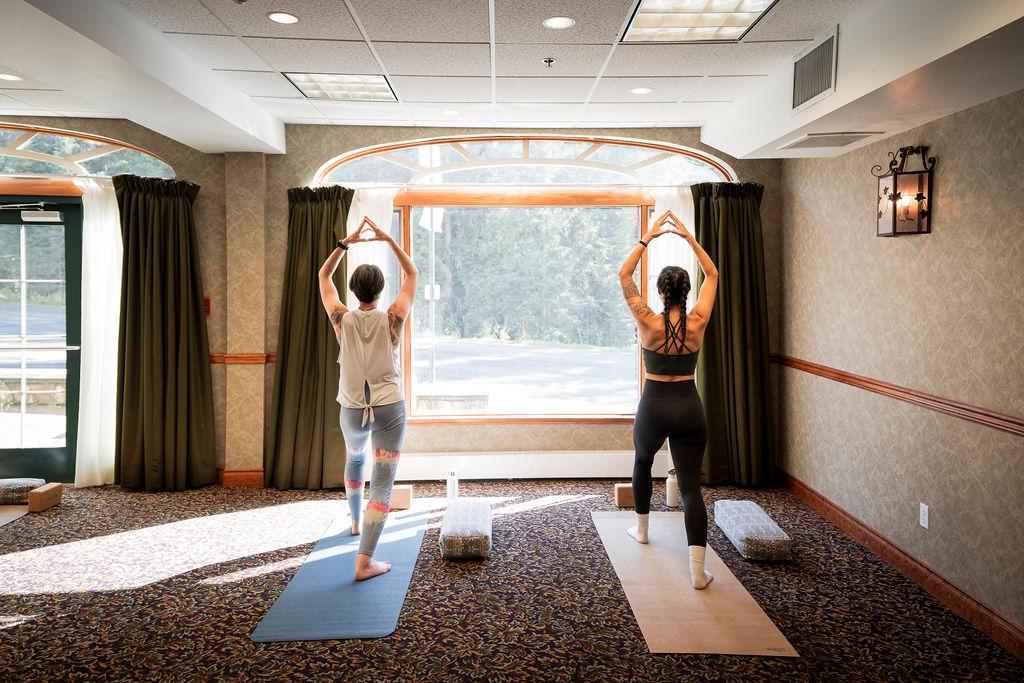 Two women practice yoga in a sunlit room, facing a large window.
