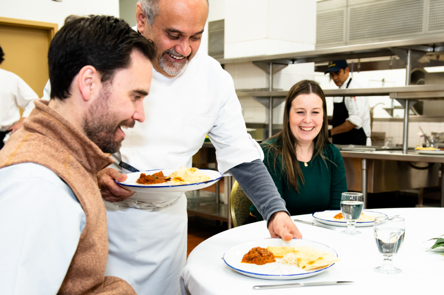 Chef serving food to smiling diners at a restaurant table.