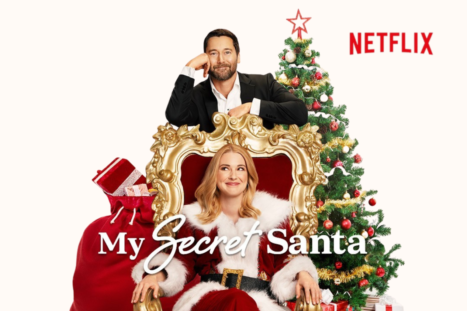 Man and woman with a Christmas tree, woman in Santa suit seated on a red and gold ornate chair, festive setting.