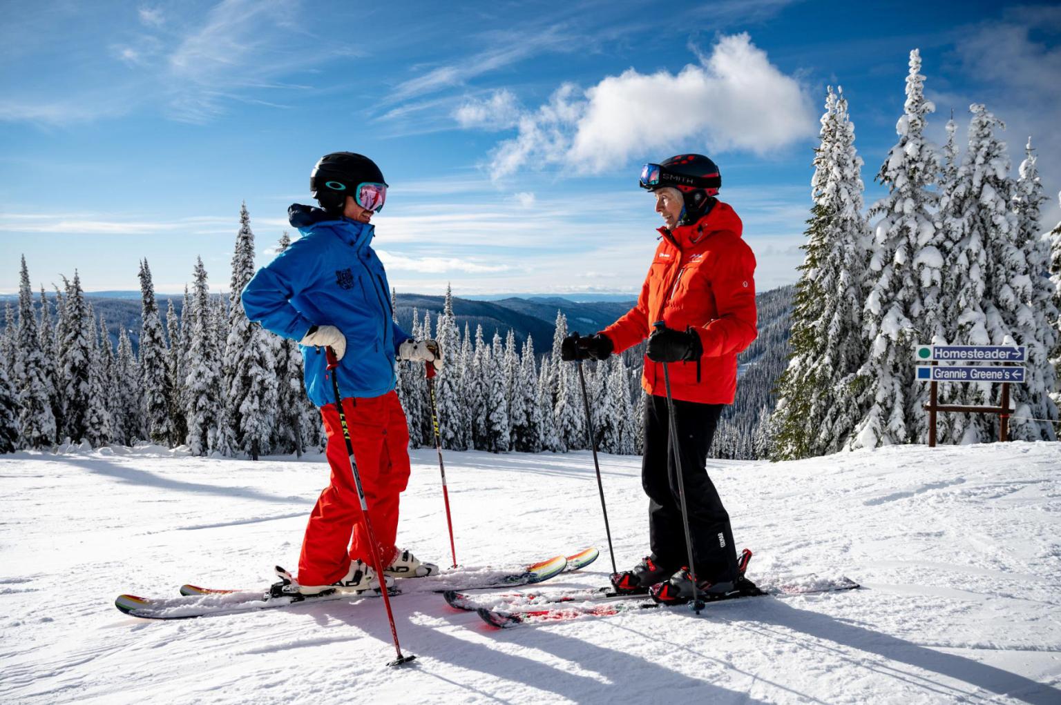 Nancy Greene chatting with a skier on snowy slope, scenic snowy trees in the background.