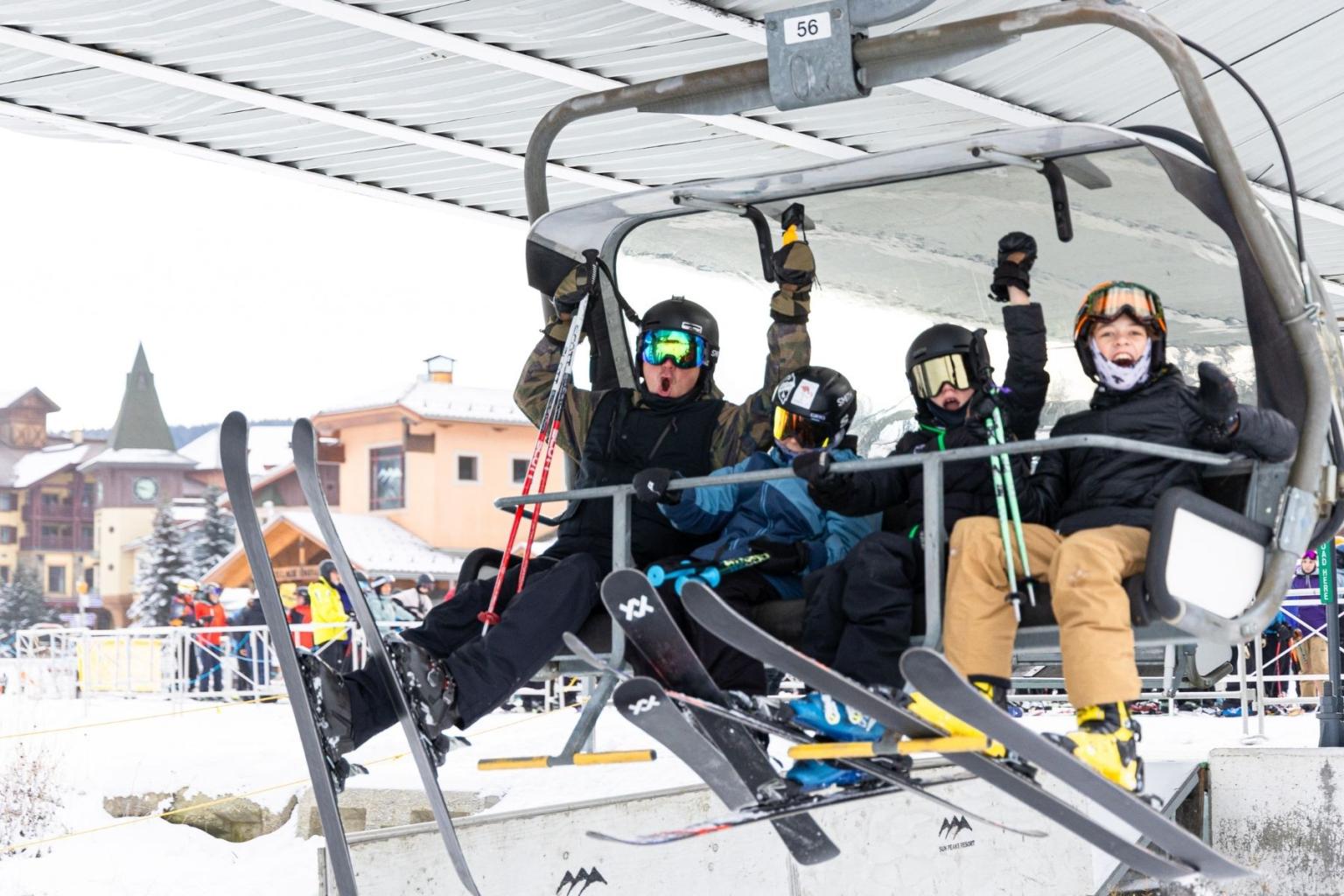 Four people on a ski lift, wearing winter gear and waving, snowy background.