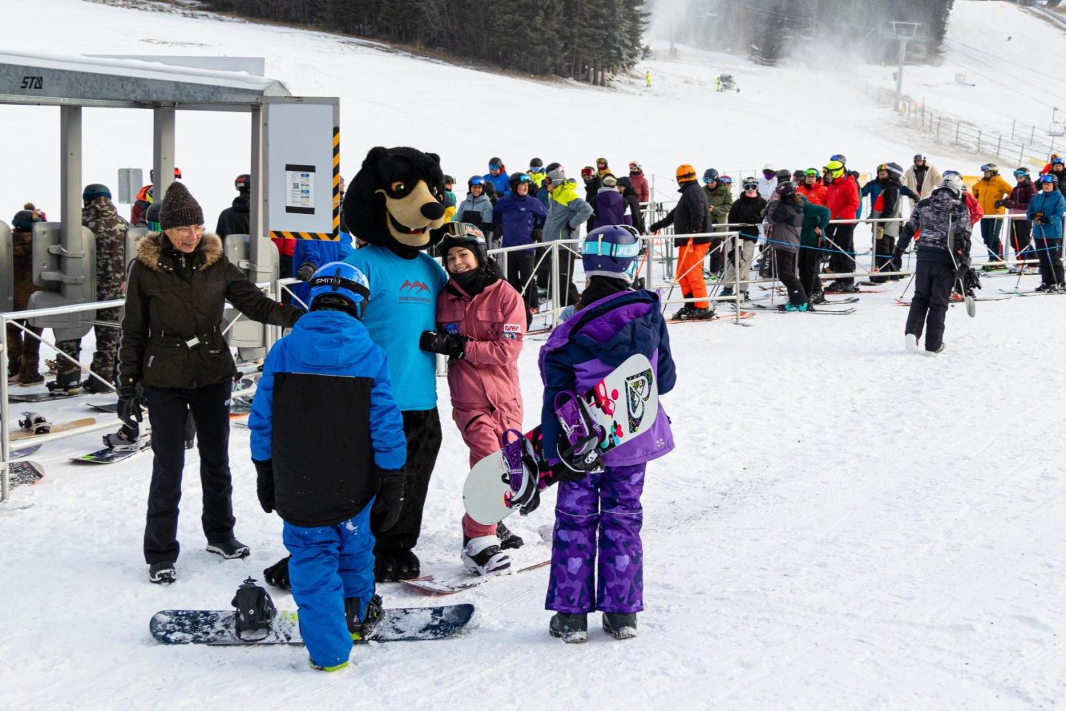 Kids with snowboards meet a costumed mascot Harry Bear on a snowy slope at Sun Peaks Resort.