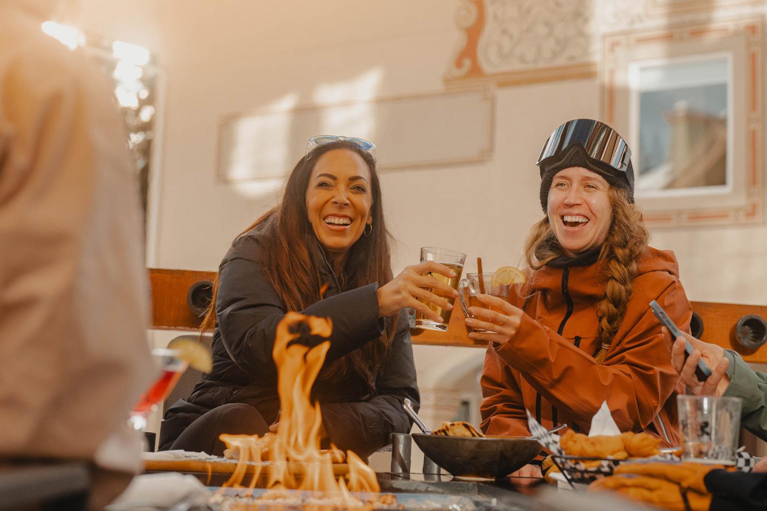Two friends toasting drinks by a cozy outdoor firepit, smiling and wearing winter clothes.