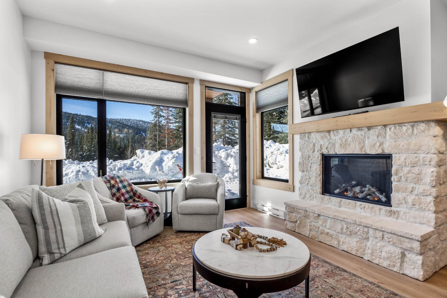 A cozy living room in the Switchback Creek condo features a sofa, a stone fireplace, and large windows with snowy views outside.