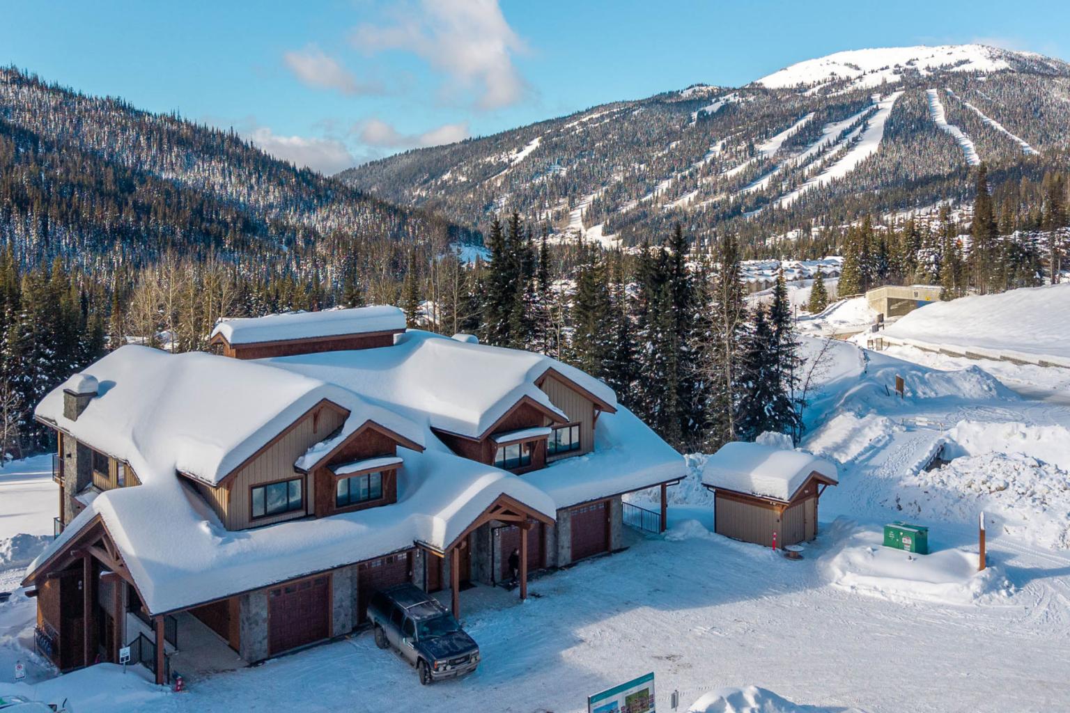 Snow-covered cabin in a mountainous winter landscape with pine trees and blue sky.