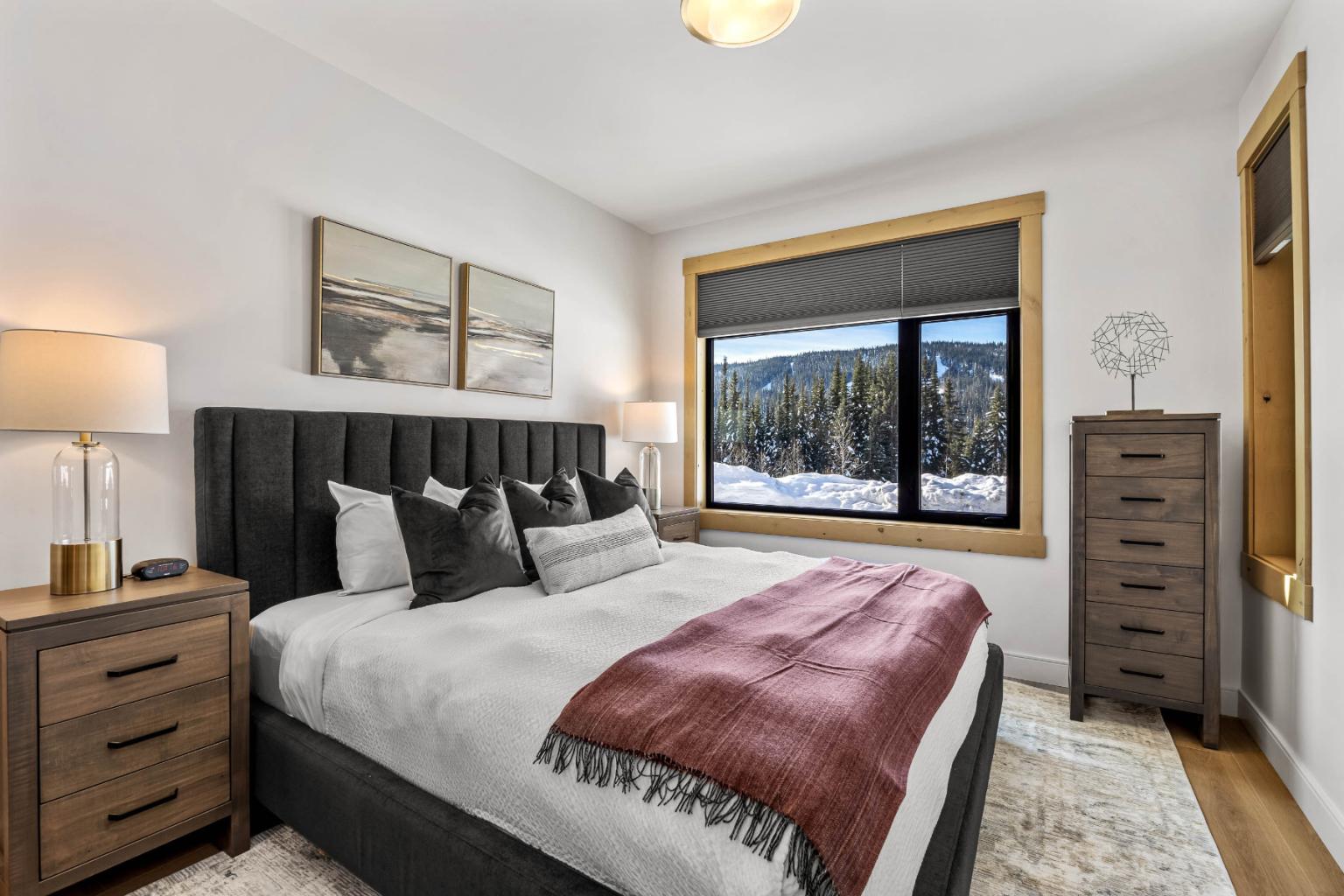 A bedroom in the Switchback Creek condo features a large bed, wooden furniture, and a window overlooking snowy mountain views.