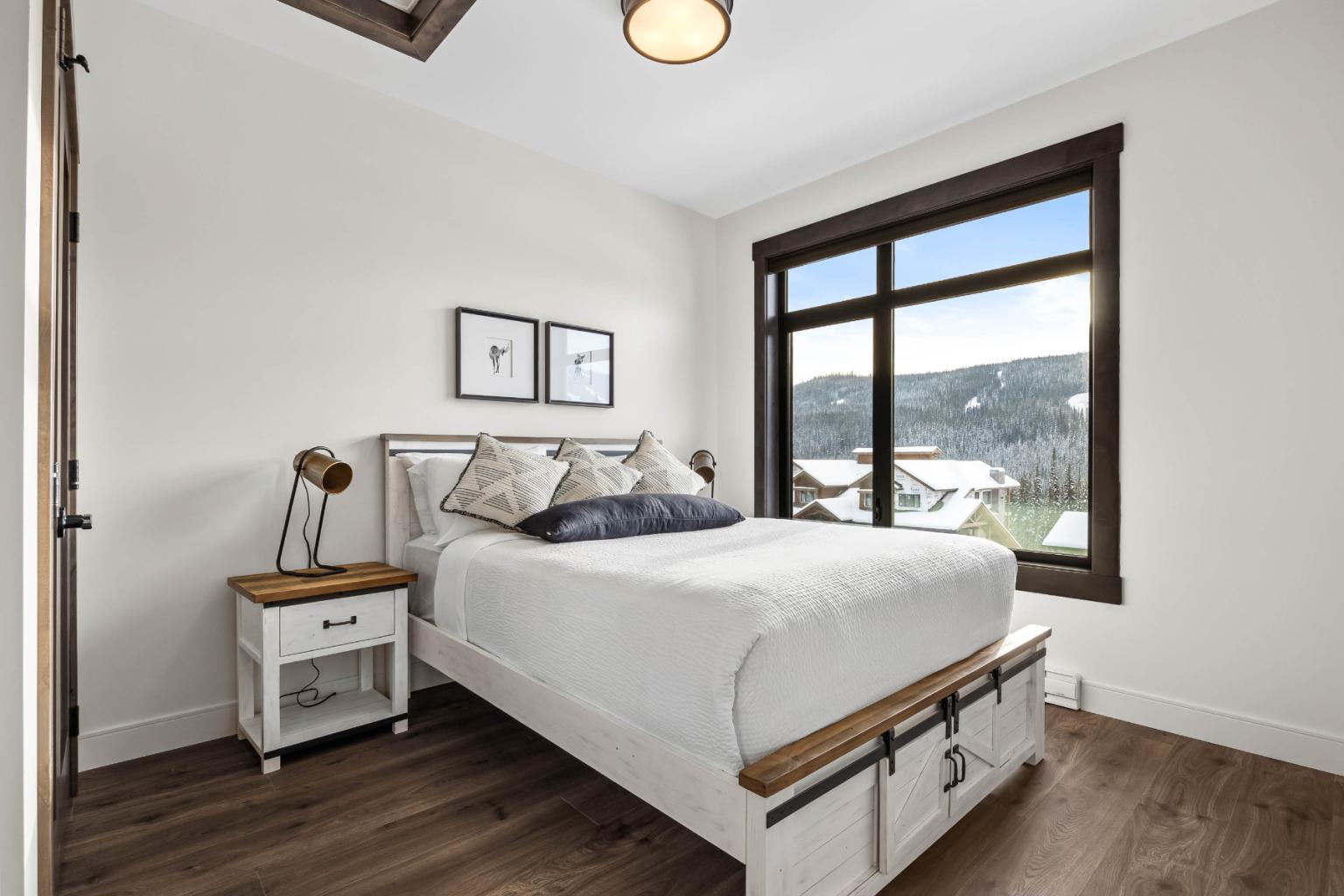 A cozy bedroom in the Switchback Creek townhome features a white bed, wooden floors, and a large window with a mountain view.