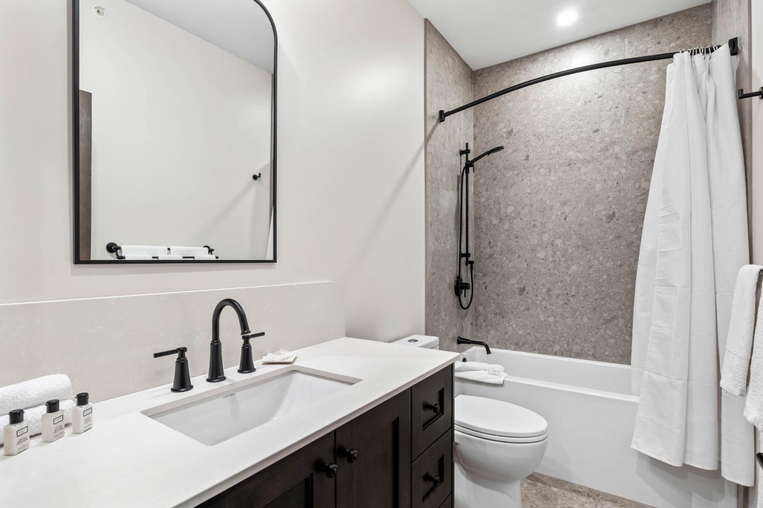 A modern bathroom in the Switchback Creek townhome features white walls, a dark wood vanity, and a shower with a curtain.