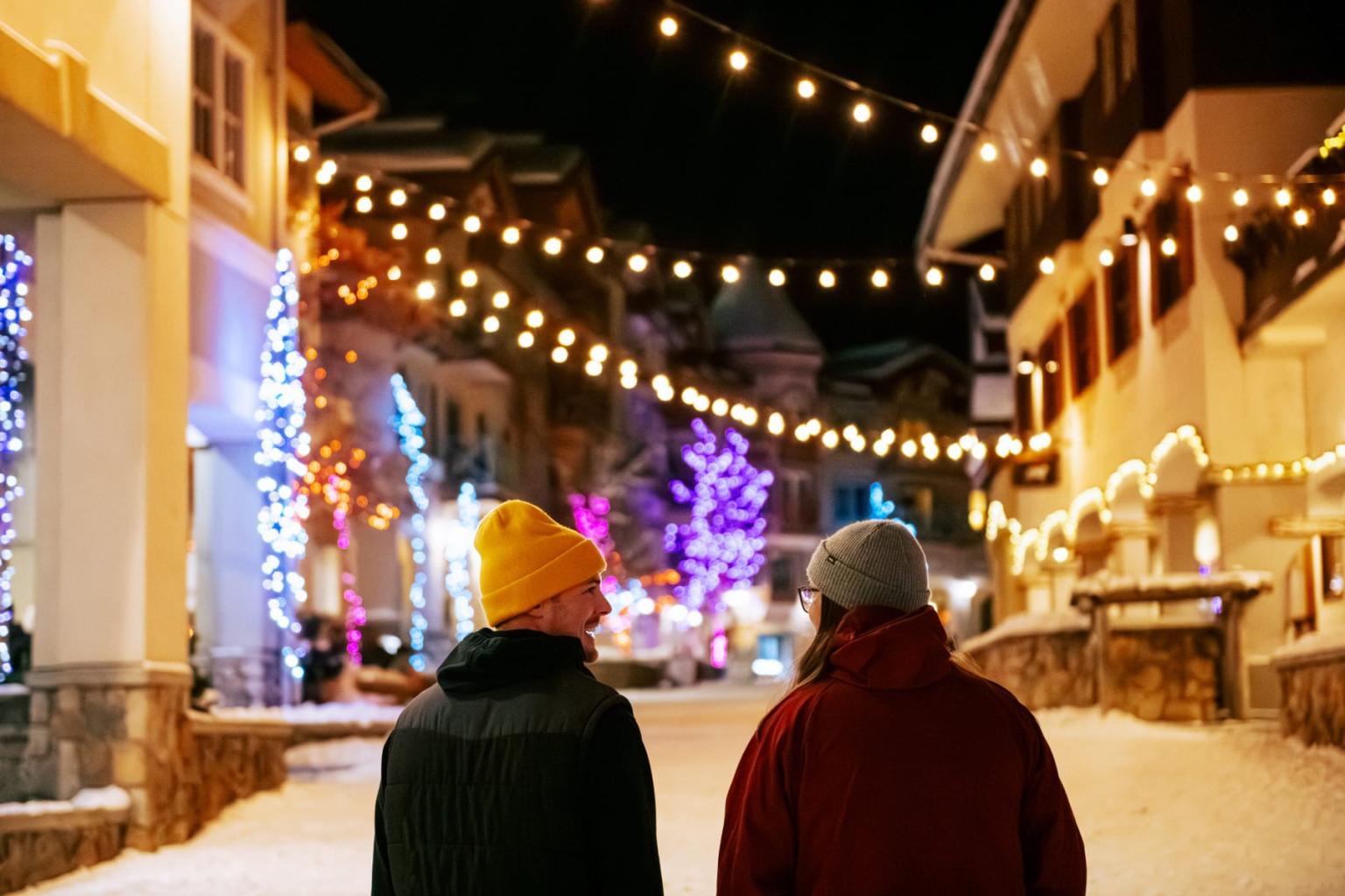 Two people in winter coats walk through a snowy, lit-up street at night.