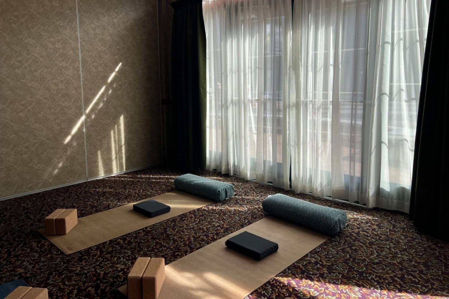 Yoga mats, blocks, and bolsters in a sunlit room with curtains drawn.