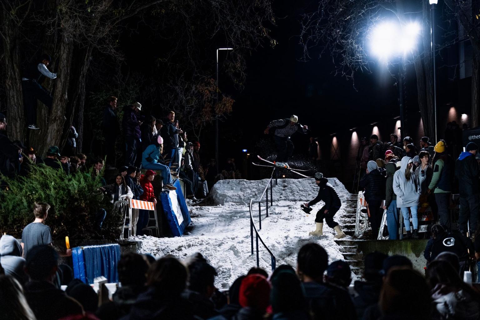 Snowboarder performs trick at night on a snowy ramp and rail, surrounded by spectators at Thompson Rivers University.