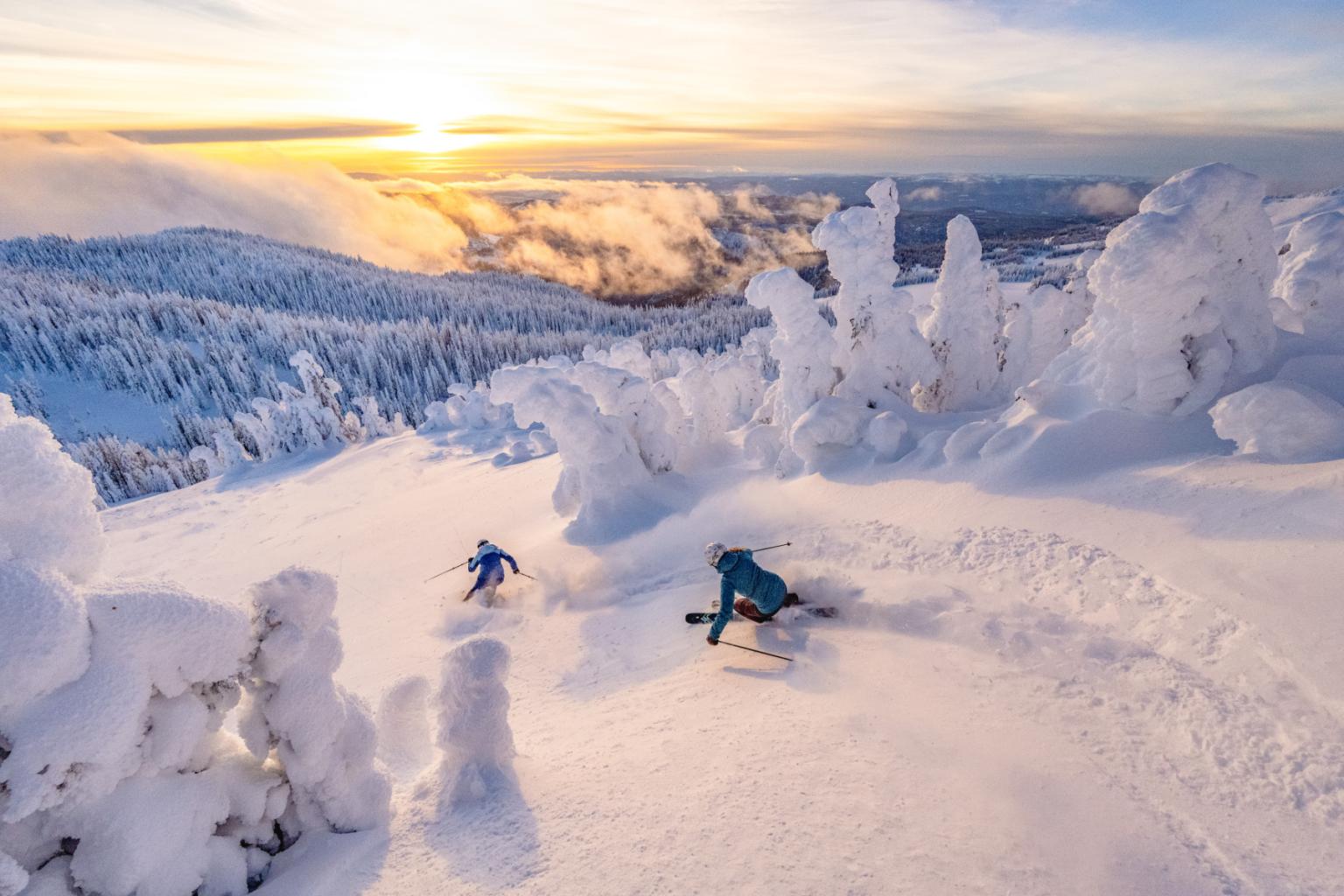 Two skiers descend a snowy slope at sunset, surrounded by frosted trees.