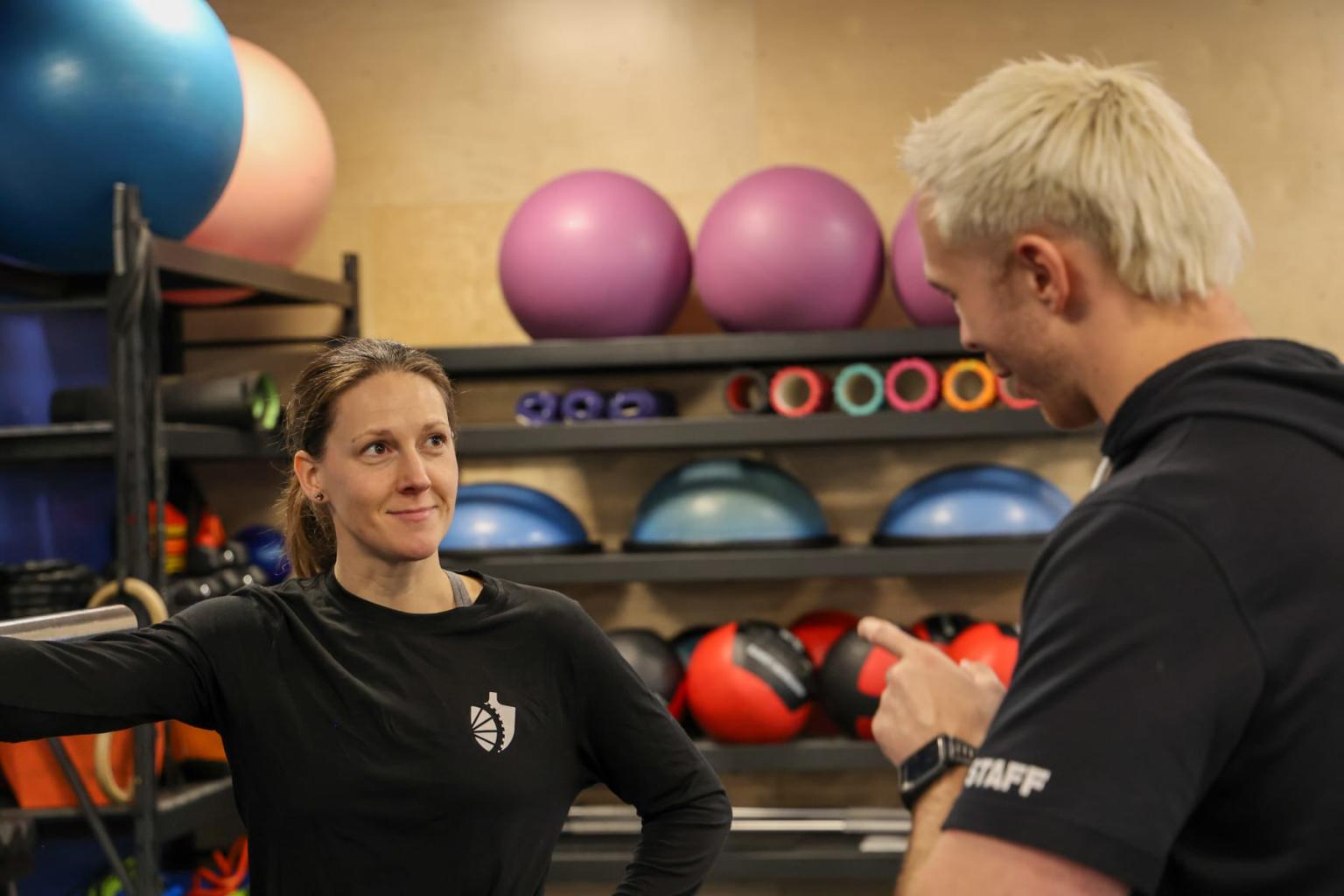 Two people talking in a gym with exercise balls on shelves.