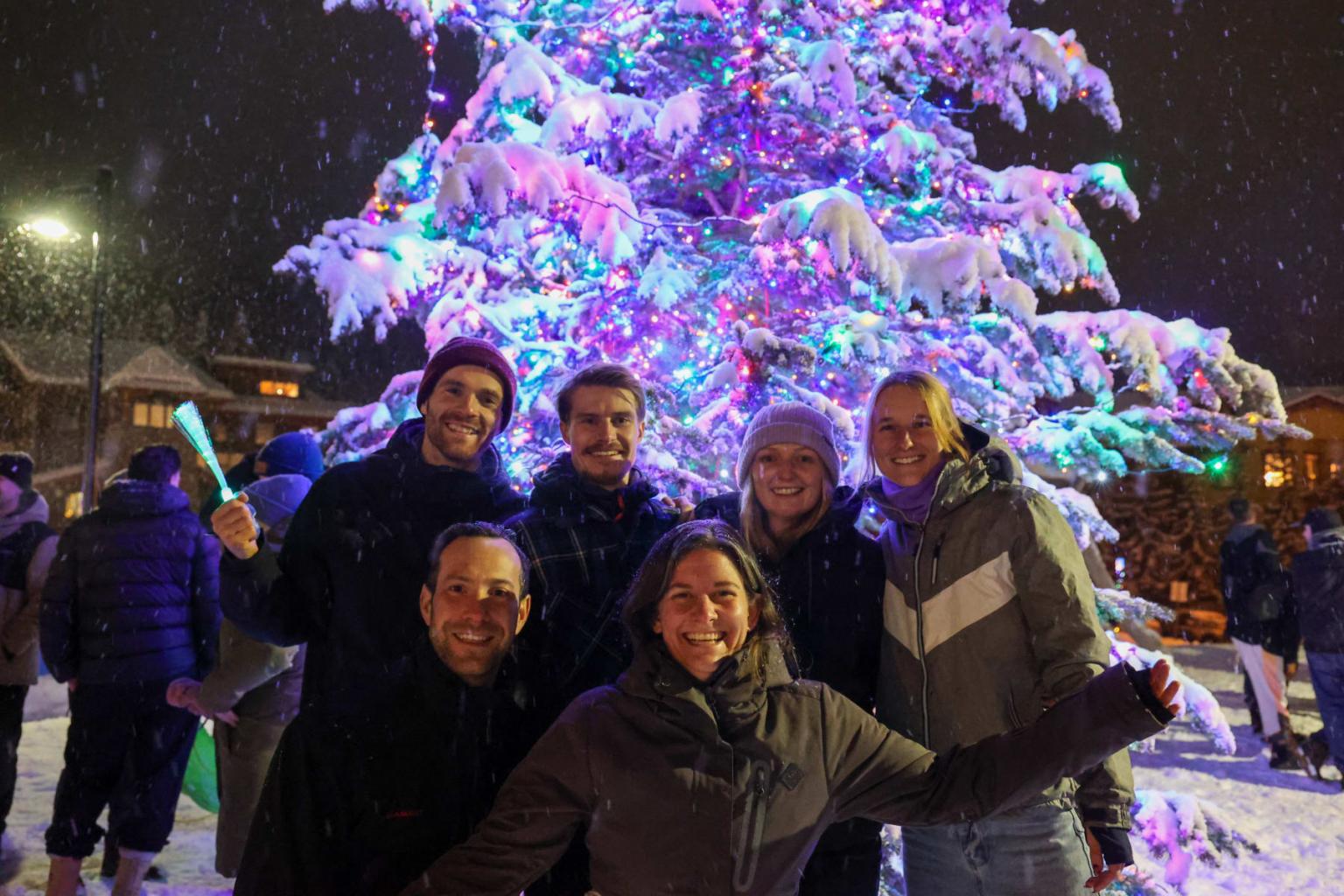 Group smiling in front of a colorful, lit Christmas tree in snowy night setting.s