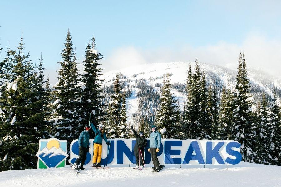 Skiers posing by a "Sun Peaks" sign, snowy mountains and trees in the background.