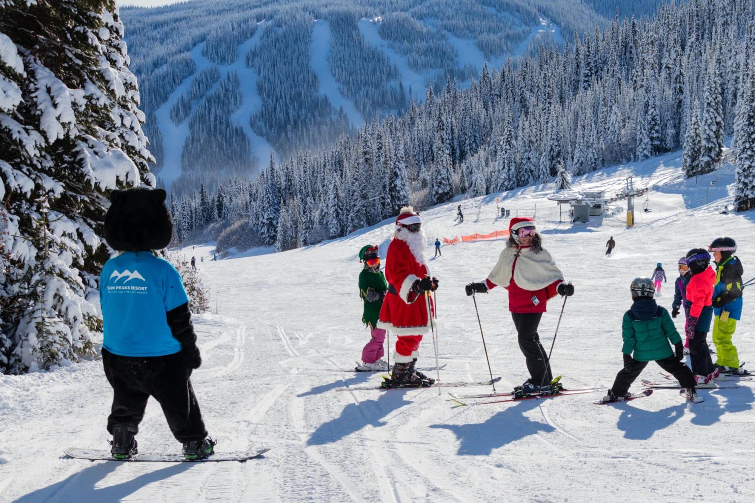 Skiers, including Santa and mascot, pause on snowy mountain slope with trees and blue sky.