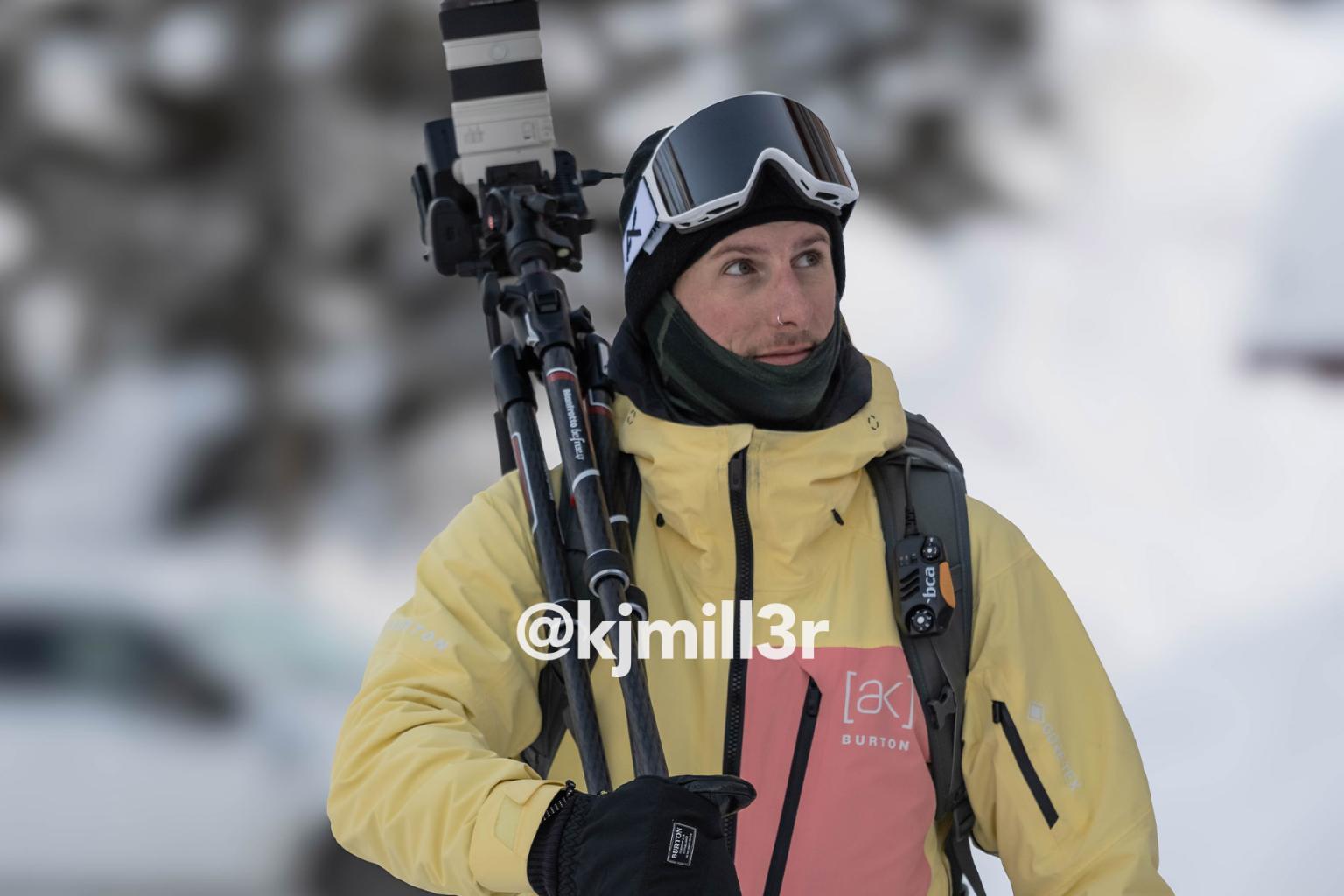 A skier in goggles and a yellow jacket carries skis on a snowy day.