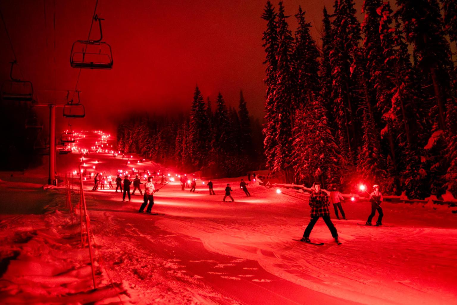 Skiers at night with red torches, surrounded by snow and trees.