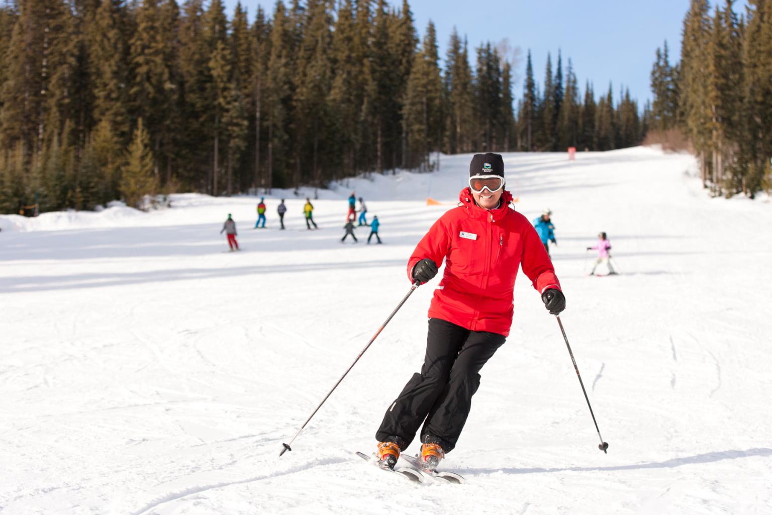 Olympic Skier Nancy Greene in red jacket glides down snowy slope, surrounded by trees and others in the distance.