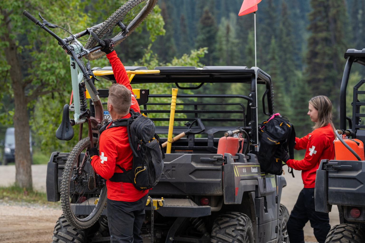 Two people in red shirts load a bike onto an ATV in a forested area.